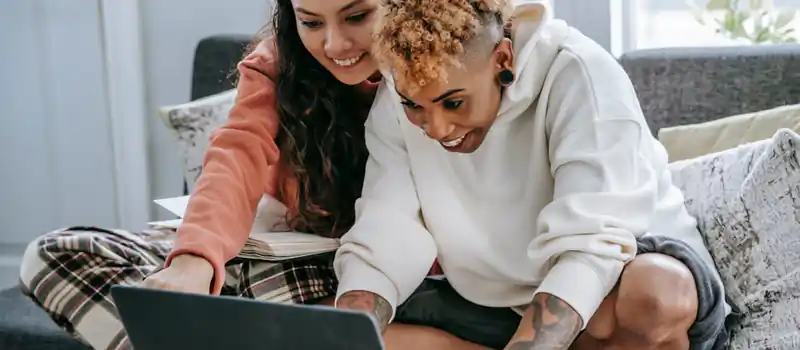 Crop cheerful multiracial women sitting with crossed legs on couch while watching netbook at home