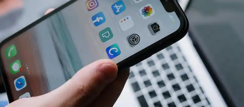 Person holding a smartphone in front of a laptop showcasing various app icons, highlighting digital connectivity.