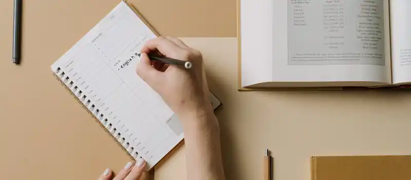 A person's hands writing in a planner at a desk with an open book nearby, conveying an organized workspace.