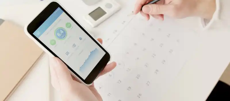Close-up of hands using a smartphone to track health stats while planning on a calendar.