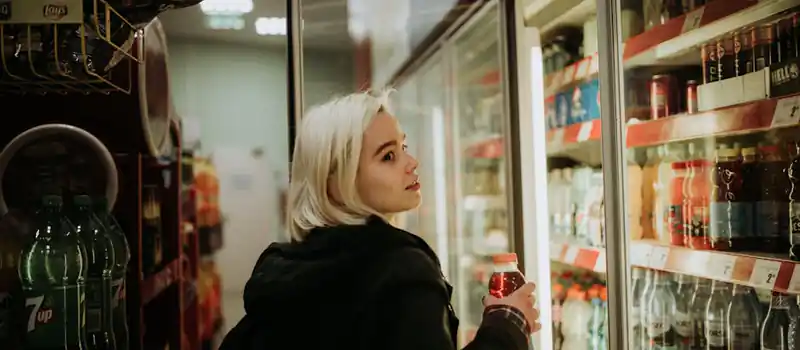 Blonde woman selecting a beverage from a refrigerated shelf at a convenience store.