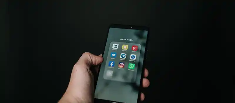 Close-up of a hand holding a smartphone displaying various social media app icons on a dark background.