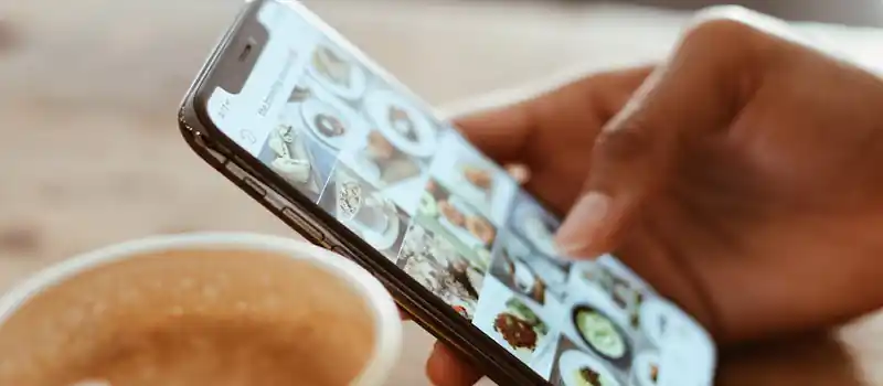 Hand browsing social media photos on a smartphone next to a cup of coffee indoors.