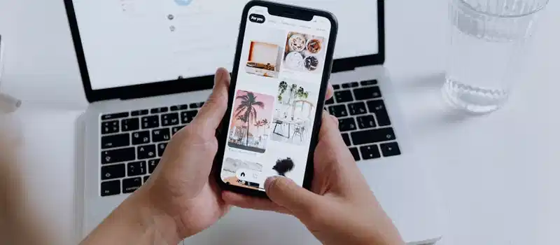 Hands holding a smartphone showcasing a gallery, with a laptop in the background and a glass of water nearby.