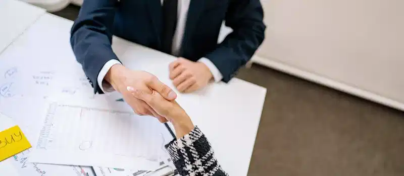 Two professionals formalizing a business agreement with a handshake over a desk.