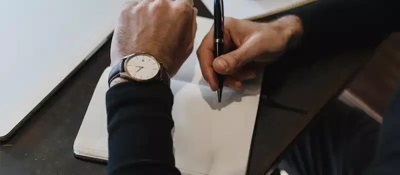 Close-up of a person writing in a notebook, wearing a watch, top view.