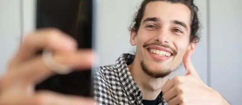 Smiling young man taking a selfie indoors with a smartphone, giving a thumbs up gesture.
