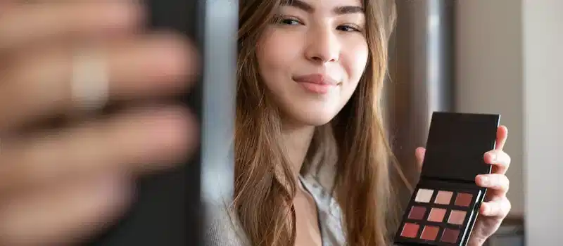 A young woman live streaming a makeup tutorial from her home office, using a smartphone.