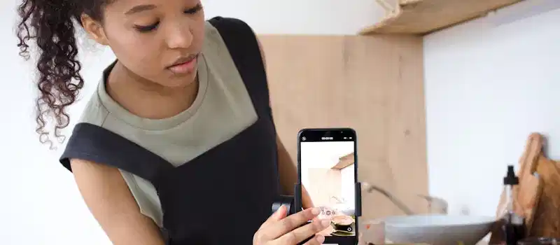A young woman filming a cooking tutorial in her kitchen using a smartphone.