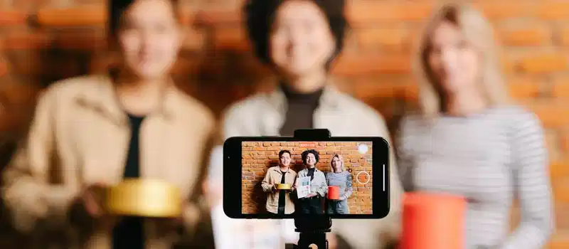 A diverse team of vloggers recording a video with a smartphone against a brick wall backdrop.