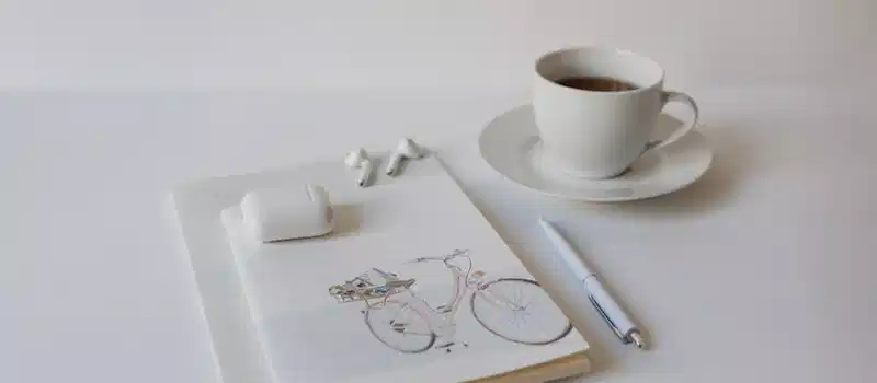 A minimalist setup with a cup of coffee, notepad, pen, and wireless earbuds on a white background.