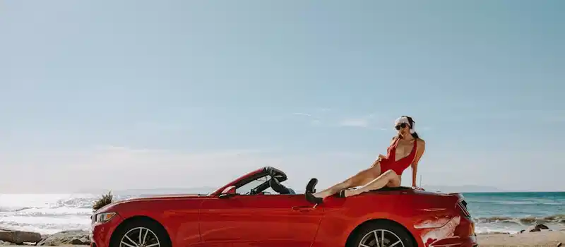 A woman in a red swimsuit lounging on a red convertible by the ocean, enjoying a sunny day.