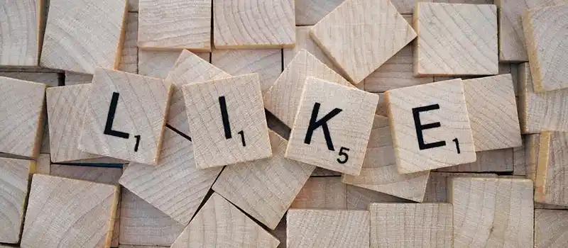 Top-down view of Scrabble tiles spelling 'LIKE' on a pile of blank wooden tiles.