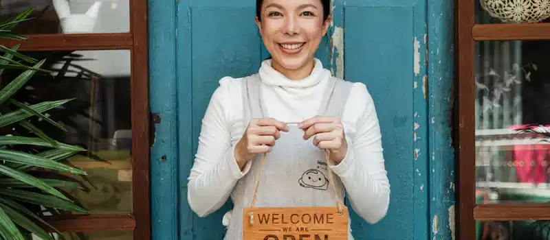 Cheerful ethnic female cafeteria owner in apron demonstrating cardboard signboard while standing near blue shabby door and windows after starting own business and looking at camera