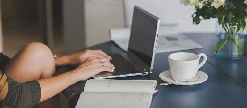 Female freelancer using laptop with coffee at home office desk, surrounded by roses and a planner.