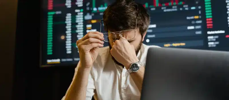 A stressed man looks at stock market data on his computer screen in an office setting.