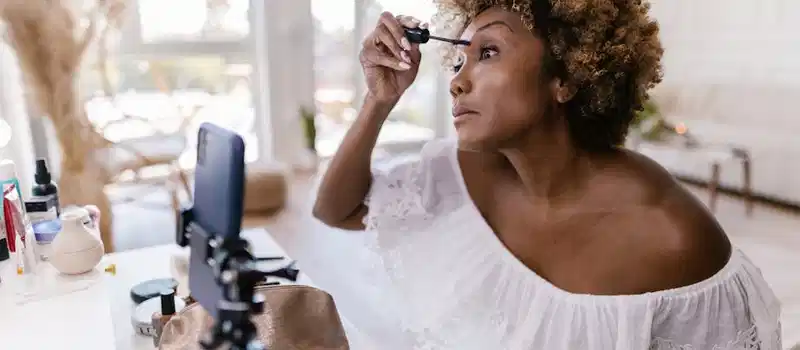 A woman applying mascara during a beauty vlog inside a bright room.