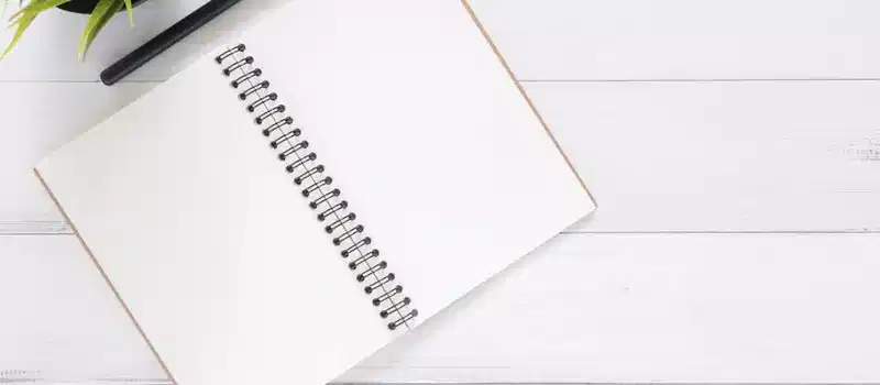 Open spiral notebook on white desk with black pen and green plant. Ideal for planning.