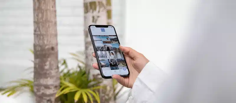 A woman using a smartphone to browse social media while standing outdoors in a tropical setting.