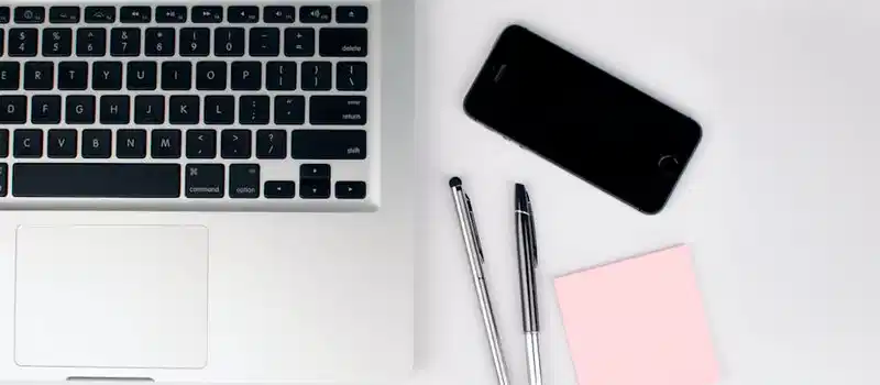 A top view of a modern workspace featuring a laptop, smartphone, pens, and a pink sticky note on a white desk.