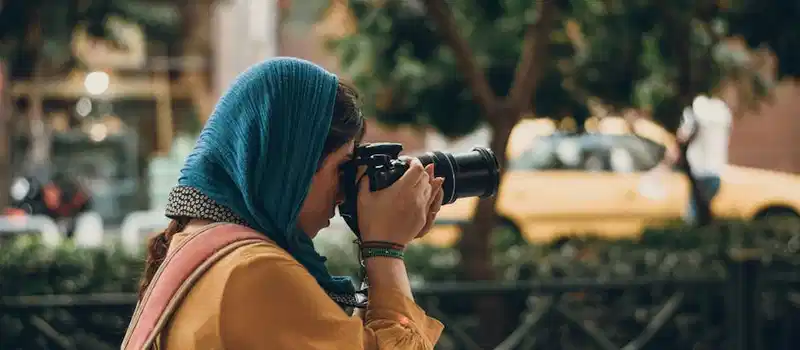 Female photographer with camera capturing street scenes, wearing casual attire and headscarf.