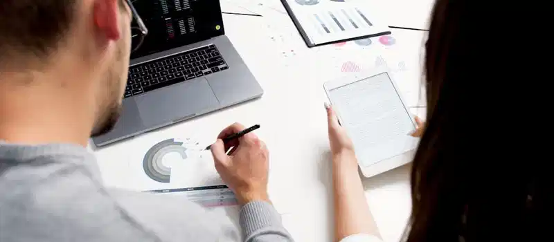 Two business professionals analyzing financial reports using a laptop and tablet in a modern office setting.