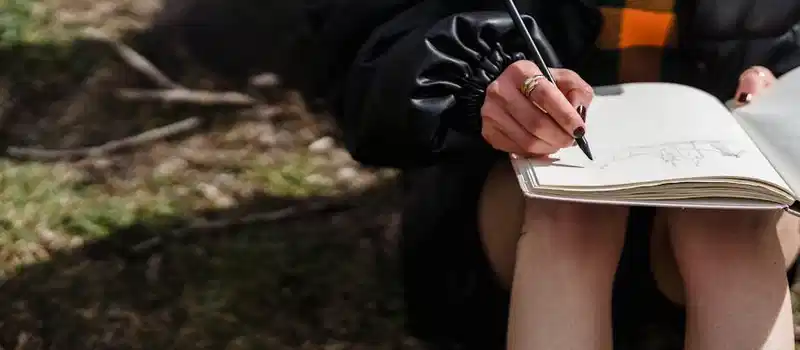 Crop unrecognizable female in black jacket drawing with pencil in workbook while sitting on meadow