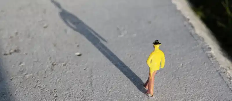 A close-up image of a toy figure in yellow clothing casting a long shadow on a concrete surface.