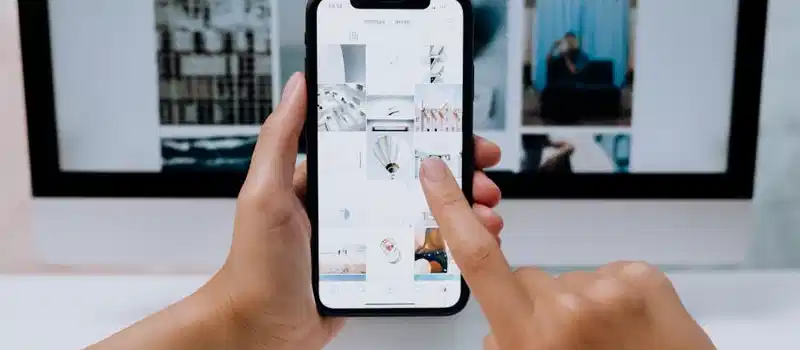Hands holding a smartphone while browsing a social media app in front of a desktop computer.