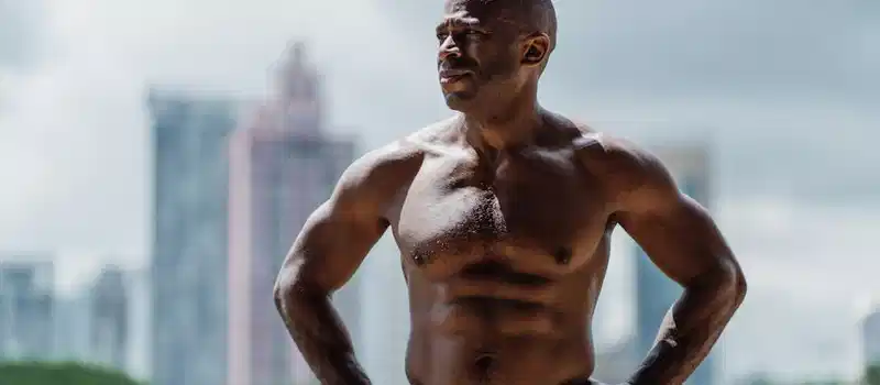 Muscular man posing shirtless outdoors against a city skyline, showcasing strength and vitality.