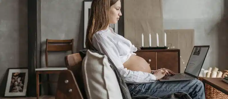 Side view of a pregnant woman using a laptop at home, emphasizing work from home lifestyle.