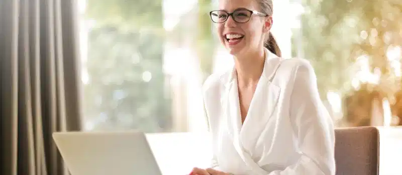Cheerful businesswoman in glasses working on a laptop, in a bright and modern office setting.