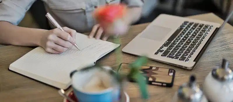 Woman writing in a notebook with a laptop and coffee cup on a desk. Ideal for workspace inspiration.