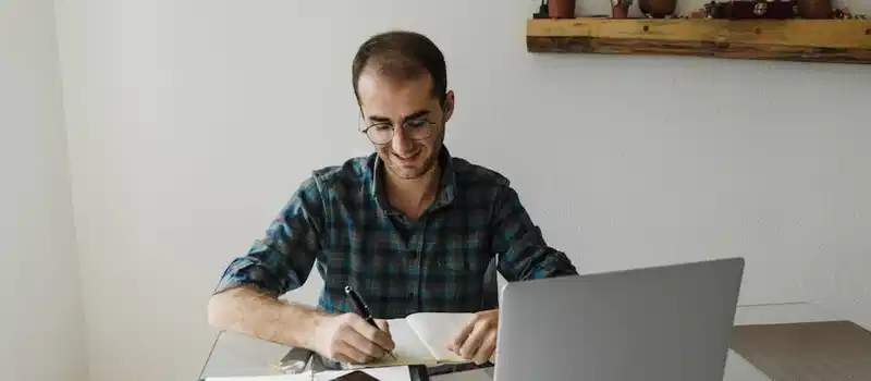 A man in a home office writing in a notebook, working with a laptop.