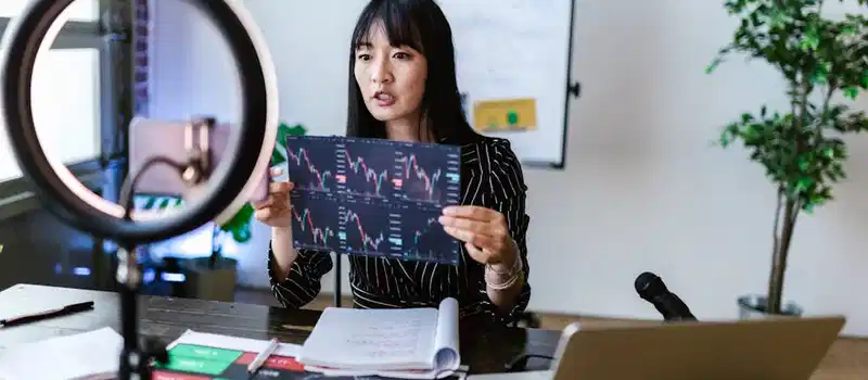 A woman vlogger discusses stock market trends using charts in a home studio.