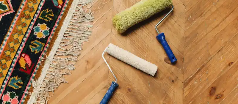 Two paint rollers on a wooden floor next to a colorful rug, symbolizing home renovation.