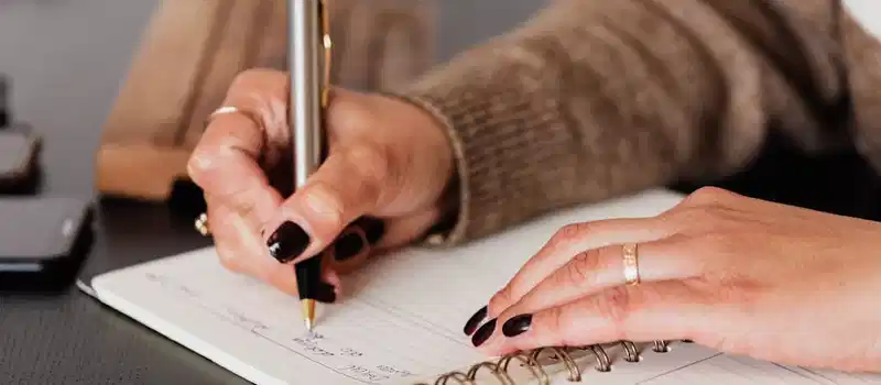 Crop unrecognizable female with stylish manicure sitting at black desk with keyboard and smartphone and taking notes with silver pen in notepad