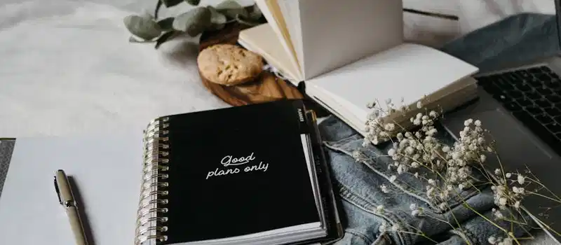 Aesthetic workspace with a notebook, pen, flowers, and laptop on a cozy surface.
