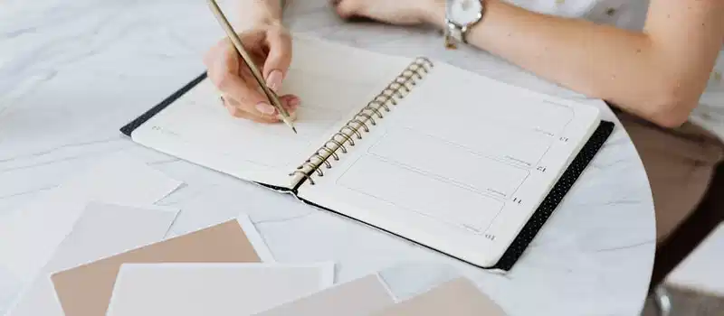 A woman writes in a planner on a marble table, surrounded by swatches. Creative and organized workspace.