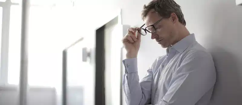Thoughtful man in a bright room holding his glasses while leaning against a wall.