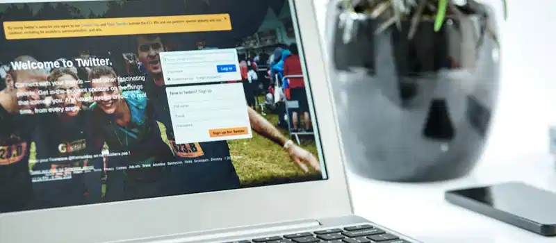 Close-up of a laptop displaying Twitter login page, accompanied by a smartphone and a potted plant in a bright workspace.