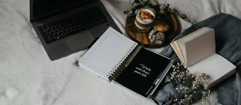 A cozy flat lay of a laptop, journals, and snacks on a bed for a relaxed work-from-home setup.