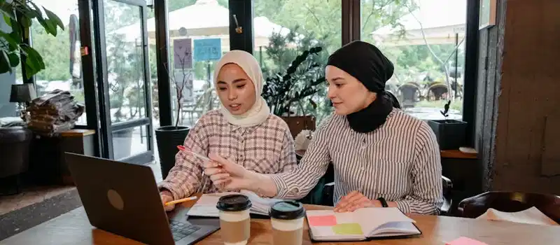 Two women in hijabs collaborating on a laptop in a cozy cafe setting, engaging in remote work.