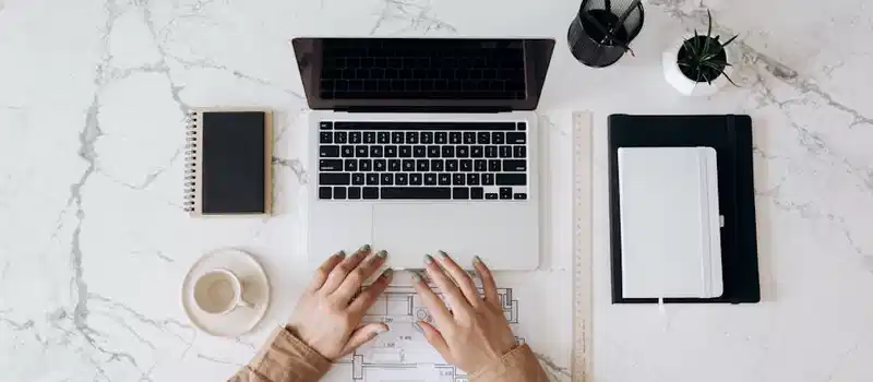 Top view of a stylish home office desk with a laptop, planner, and coffee cup, showing hands on a blueprint.