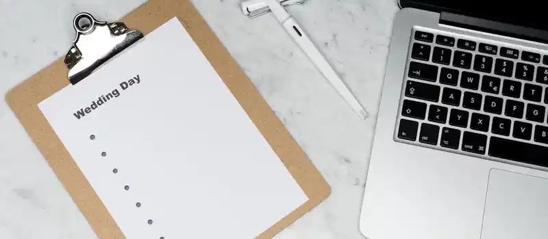 Flat lay of a wedding planning setting with clipboard, laptop, and pen.