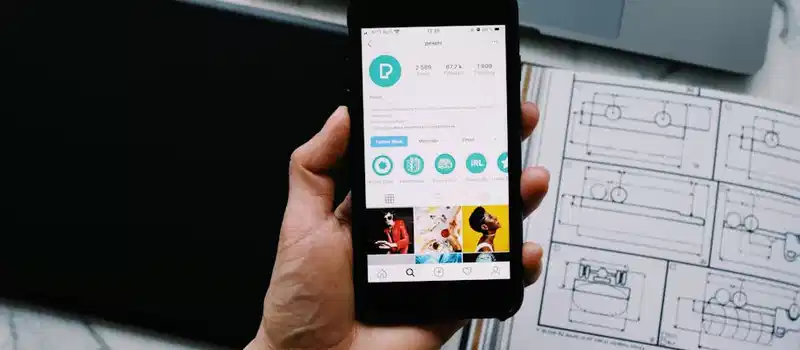 Hand holding smartphone displaying social media profile with tech gadgets on desk.