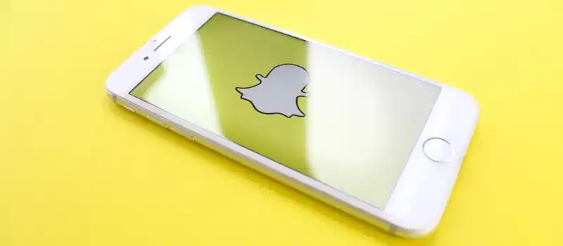 White smartphone displaying Snapchat logo on a bright yellow background, angled view.