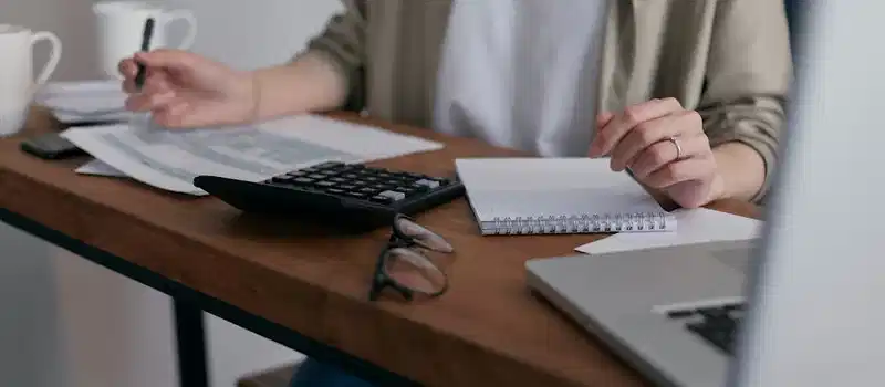 A woman manages finances at home, using a laptop and calculator on a wooden desk.