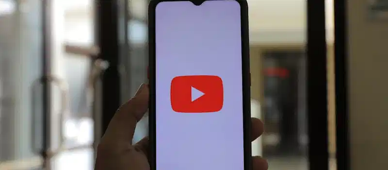 A hand holding a smartphone displaying the YouTube app indoors.