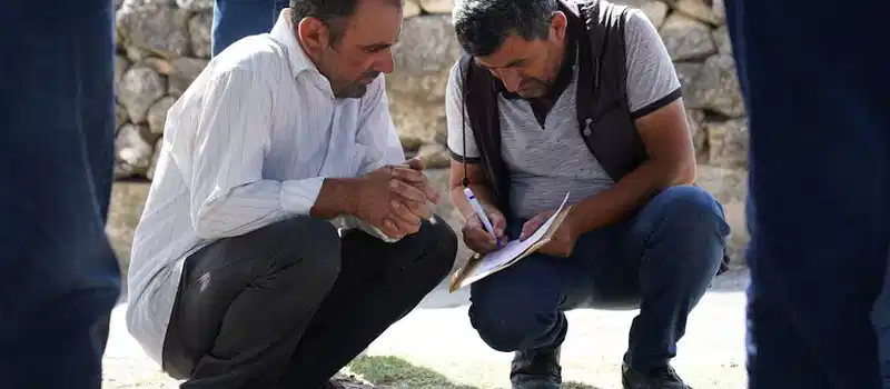 Two men squatting and analyzing paperwork outdoors in a sunny setting.